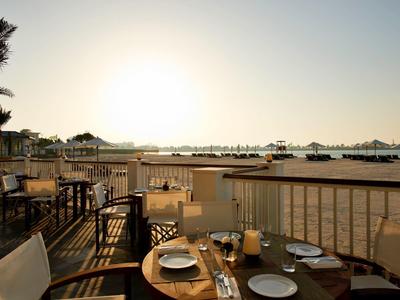 Terrassenrestaurant mit gedeckten Tischen, Holzmöbeln und Sonnenuntergang am Strand im Hintergrund.