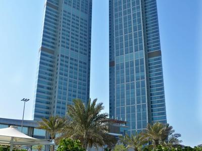 Due moderni grattacieli con facciate in vetro collegati da un ponte, cielo azzurro e piscina.