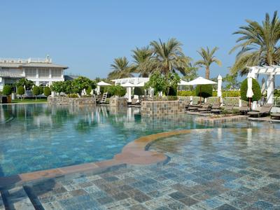 Großer Pool mit blauer Wasserfläche, umgeben von Palmen, Liegen und weißer Architektur bei klarem Himmel.