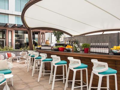 Bar all'aperto con sgabelli bianchi e cuscini blu sotto un grande tendalino.