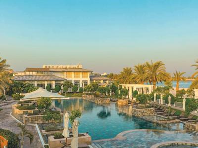 Hotel di lusso con grande piscina, palme e ombrelloni sotto un cielo sereno.