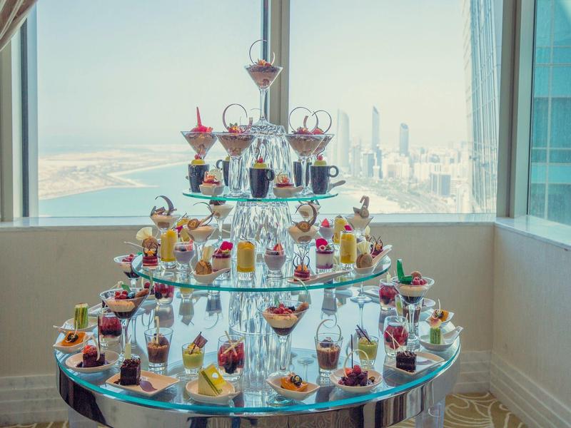 Estante de vidrio de varios niveles con diversos postres y cócteles frente a ventanas panorámicas con vista al mar.