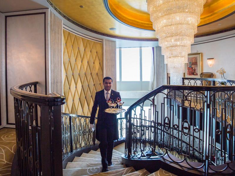 Un camarero lleva una bandeja en una elegante escalera de caracol con candelabro y grandes ventanas.