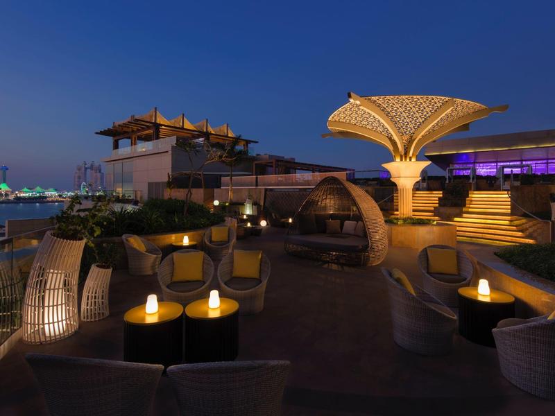 Terraza en la azotea iluminada por la noche con áreas de descanso, lámparas decorativas y vista a la ciudad.