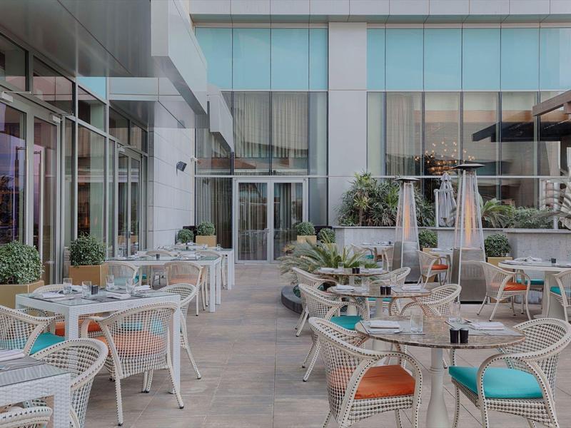 Área exterior moderna de un hotel con sillas y mesas blancas, decorada con plantas.