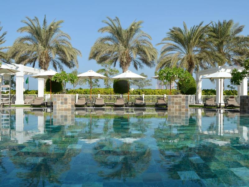Una piscina tranquila con agua reflectante, rodeada de palmeras y sombrillas blancas.