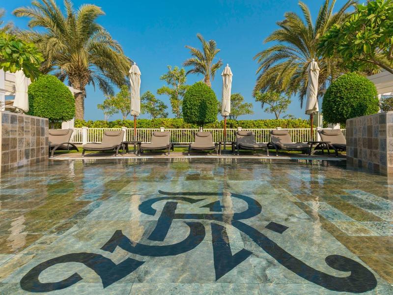 Piscina lujosa con agua clara, palmeras y tumbonas bajo un cielo azul.
