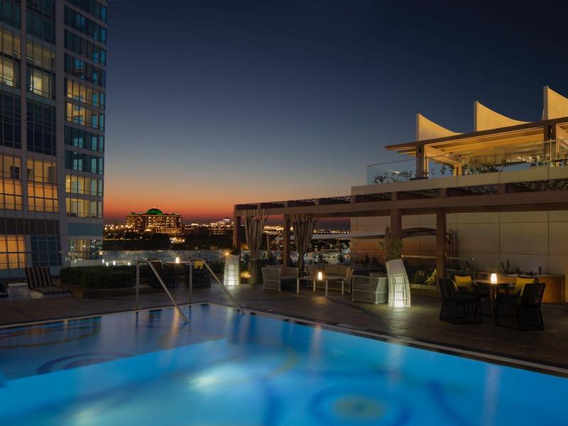 Terraza en la azotea con piscina iluminada y áreas de descanso al atardecer en un hotel.