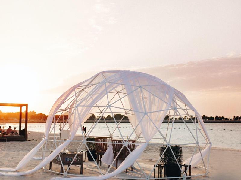 Carpa de cúpula transparente en la playa con asientos al atardecer junto al agua.