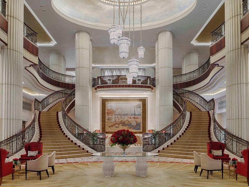 Elegante vestíbulo de hotel con escalera doble, columnas altas y mobiliario rojo.