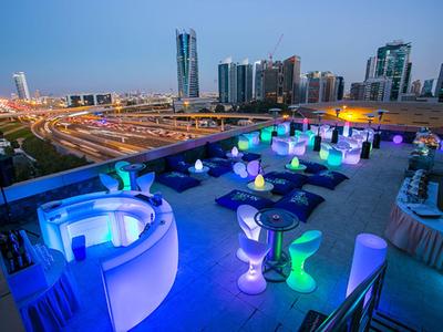 Rooftop terrace with colorful lighting, seating, and view of modern skyscrapers at night.
