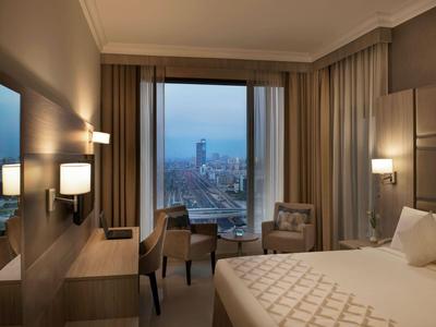 Modern hotel room with double bed, desk, chairs, and large window view of a city.