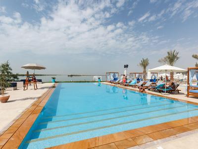 Modern hotel pool with lounge chairs and sea view under a cloudy sky.