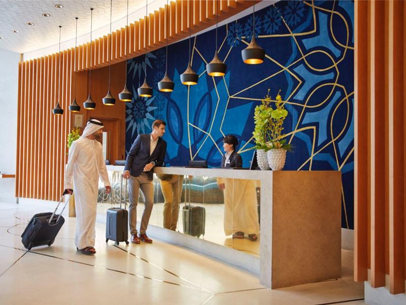 Moderne Hotellobby mit blauer Wand, Holzpaneelen und Empfangstresen, Gäste mit Koffern stehen dran.