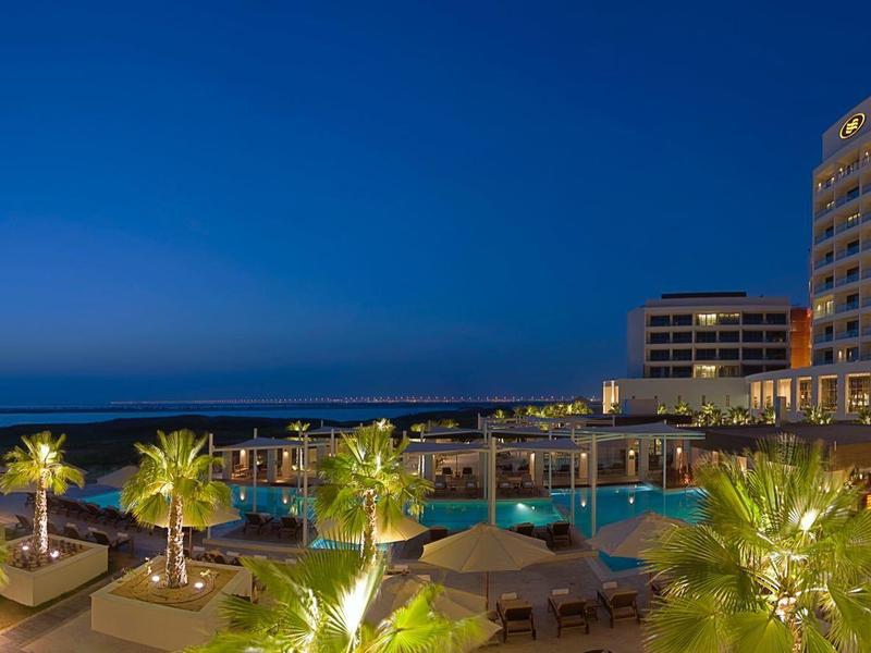Luxuriöses Hotel mit beleuchtetem Pool und Palmen bei Dämmerung am Meerufer.