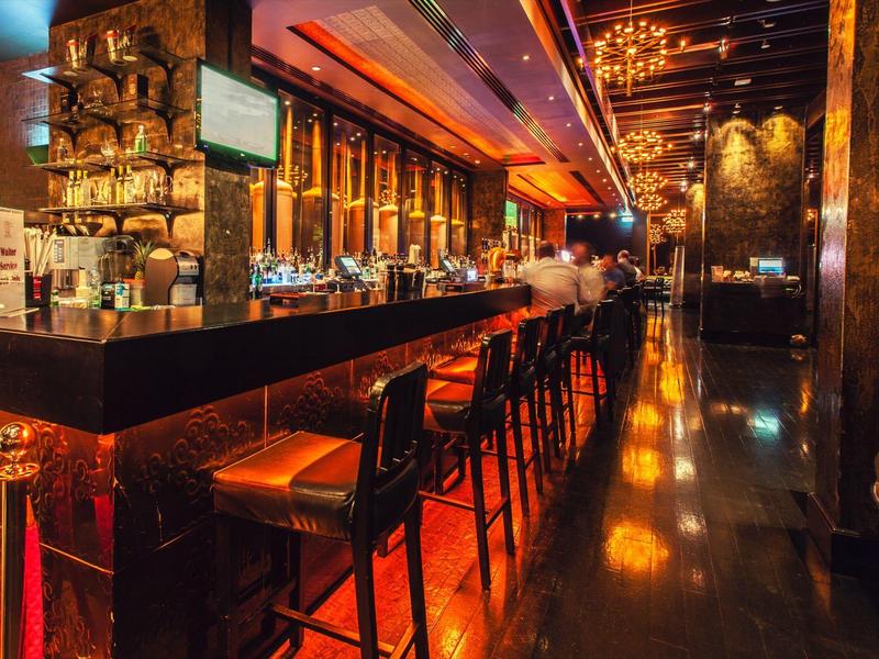 Moderne Bar mit dunklem Holz, oranger Beleuchtung und hohen Barhockern an langer Theke.