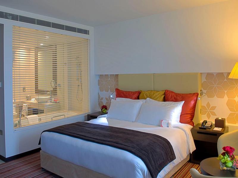 Gemütliches Schlafzimmer mit großem Bett, gelber Stehlampe und angrenzendem Badezimmer hinter Glaswand.