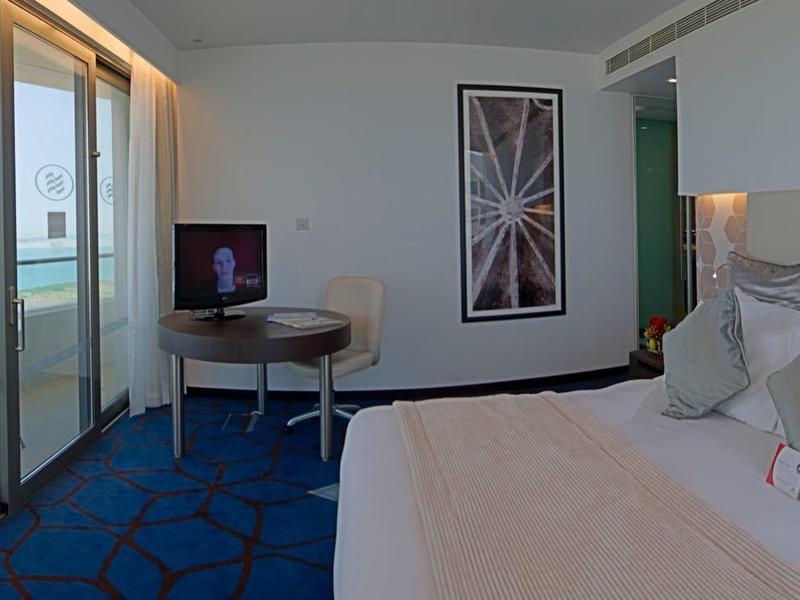 Modernes Hotelzimmer mit großem Bett, blauem Teppich und Balkon mit Meerblick.