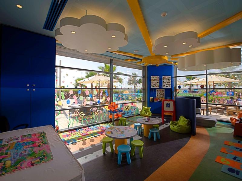 Ein heller, farbenfroher Kinderspielraum mit kleinen Tischen, Sitzkissen und großen Fenstern.