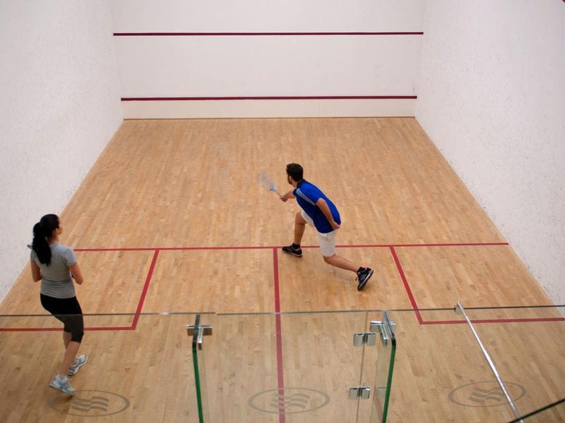 Zwei Personen spielen Squash in einem hellen, kleinen Hallenraum mit Holzboden und roten Linien.