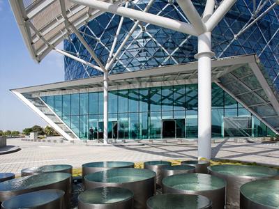 Entrada de edificio moderno con gran marquesina de vidrio y esculturas redondas de bronce en primer plano.