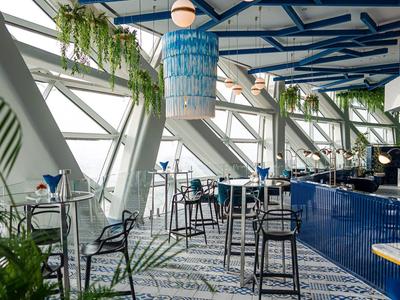 Área de comedor moderna con barra azul, ventanas altas y plantas en un hotel.