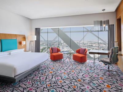 Habitación de hotel moderna con cama grande, alfombra colorida y ventana panorámica con vista a la ciudad.