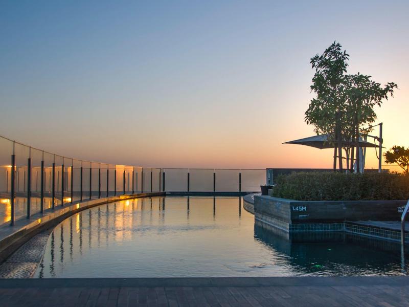 Piscina en la azotea con barandilla de vidrio y árbol al atardecer sobre una ciudad.
