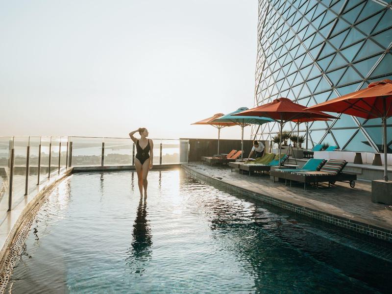 Mujer en piscina infinita con vista a la ciudad junto a edificio moderno de vidrio y sombrillas rojas.