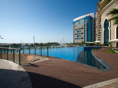 Blick auf einen modernen Infinity-Pool mit Holzdeck und Hochhaus im Hintergrund unter klarem Himmel.