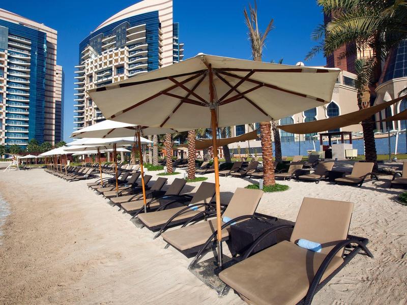 Strand mit Sonnenschirmen, Liegestühlen und modernen Hochhäusern im Hintergrund bei sonnigem Wetter.