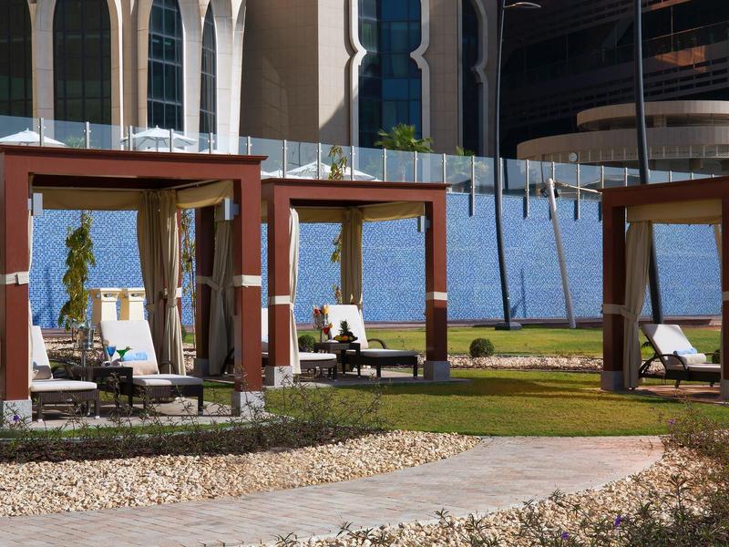 Drei hölzerne Pavillons mit weißen Liegen auf grünem Rasen vor modernen Gebäuden bei Sonnenschein.