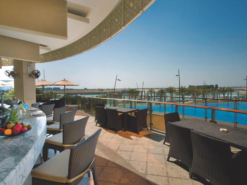Terrasse mit Sitzgruppen, Sonnenschirmen und Blick auf Meer unter klarem blauen Himmel.