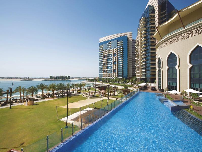 Moderner Hotelpool mit klarblauem Wasser, grüner Wiese, Palmen und hohen Gebäuden am Meer.