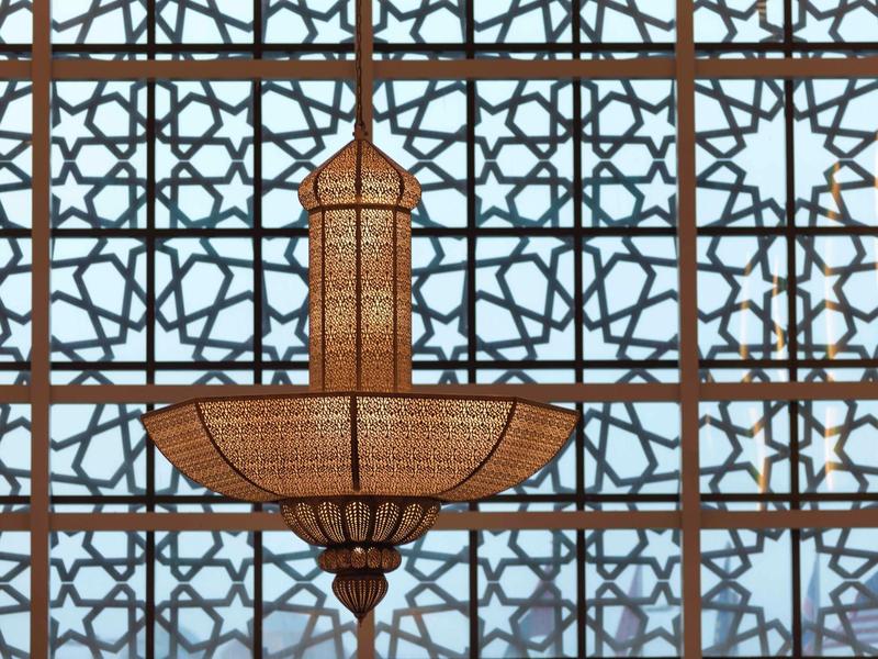 Großer, goldener Kronleuchter mit geometrischem Muster vor einem Fenster mit arabischem Gitter.