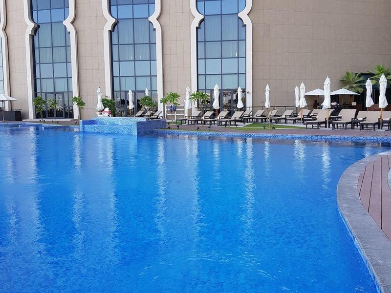 Großes blauen Schwimmbad vor einem Gebäude mit hohen, spitzbogenförmigen Fenstern und Liegestühlen.