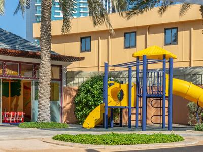 Aire de jeux pour enfants avec toboggan et structure d'escalade devant un bâtiment d'hôtel avec des palmiers.