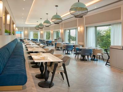 Restaurant moderne avec banquettes bleues, tables rondes et grandes fenêtres donnant sur la verdure.