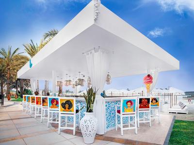 Espace bar extérieur moderne avec chaises blanches et œuvres colorées sous un auvent blanc.
