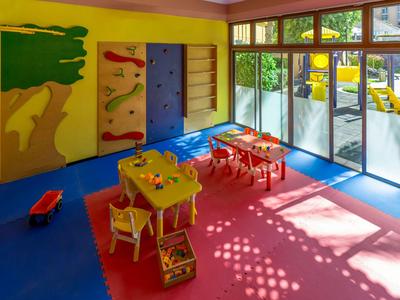Salle de jeux colorée avec tables, chaises et grandes baies vitrées donnant sur l'extérieur.