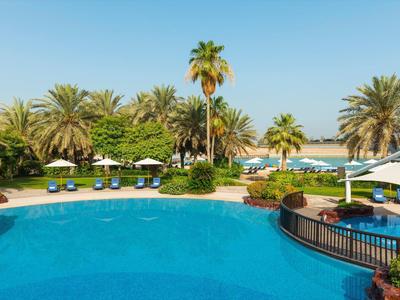 Large outdoor pool with sun loungers, umbrellas, and palm trees in a tropical hotel setting.