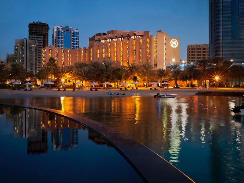 Illuminated hotel building with pool and tall buildings in the background at dusk.