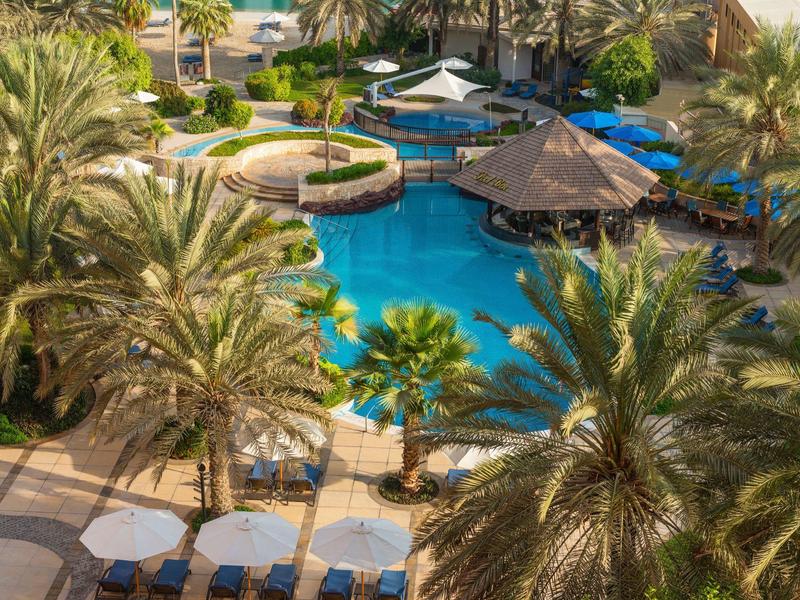 Hotel pool with palm trees, sun umbrellas, and tropical surroundings in clear weather.