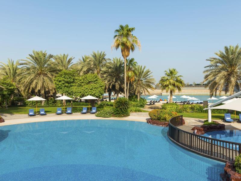 Round hotel pool with palm trees, lounge chairs, and umbrellas under clear sky.