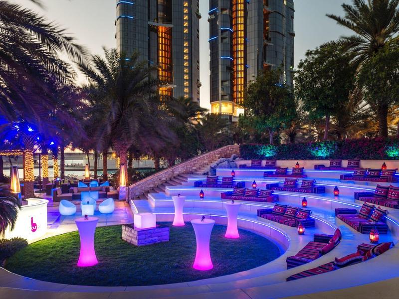 Night view of a stylish outdoor lounge with colorful glowing tables and modern skyscrapers in the background.