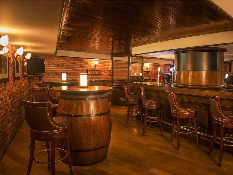 Cozy hotel bar with wooden barrel tables and comfortable bar stools in warm lighting.