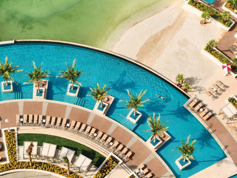 Luchtfoto van een halfcirkelvormig zwembad met ligstoelen en palmbomen aan het strand