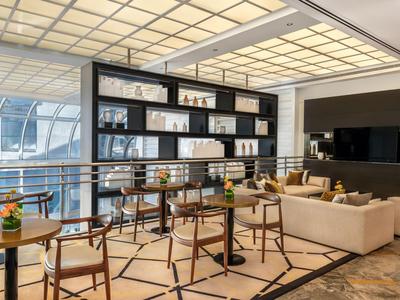 Moderní hotelová lounge s stolky, židlemi, pohovkami a velkým regálem pod jasným stropním osvětlením.