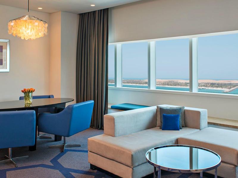 Modern eingerichtetes Hotelzimmer mit Sofa, Esstisch und großer Fensterfront mit Meerblick.