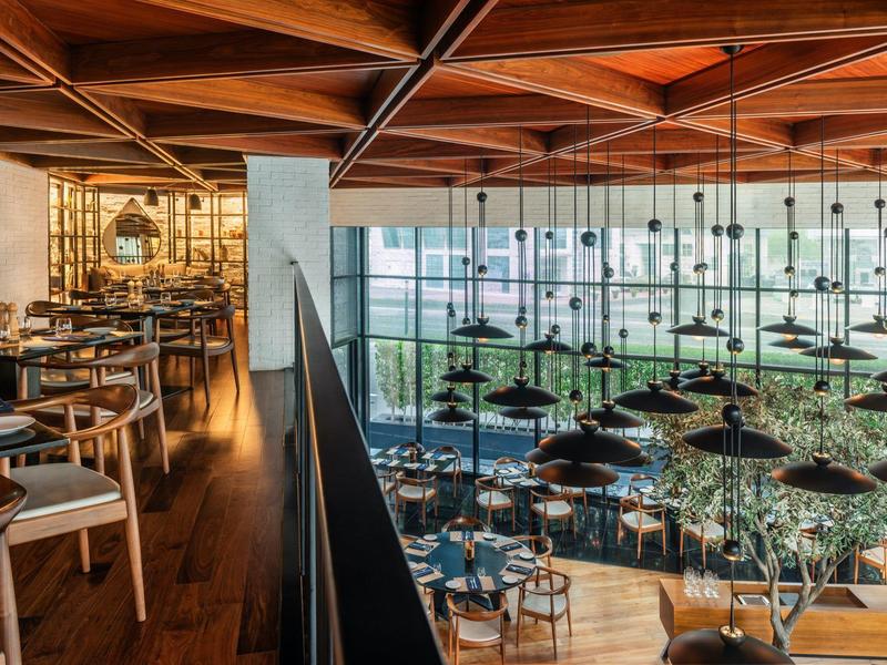 Modernes Restaurant mit Holzdecke, großen Fenstern und eleganten Tischen und Stühlen in hellem Licht.